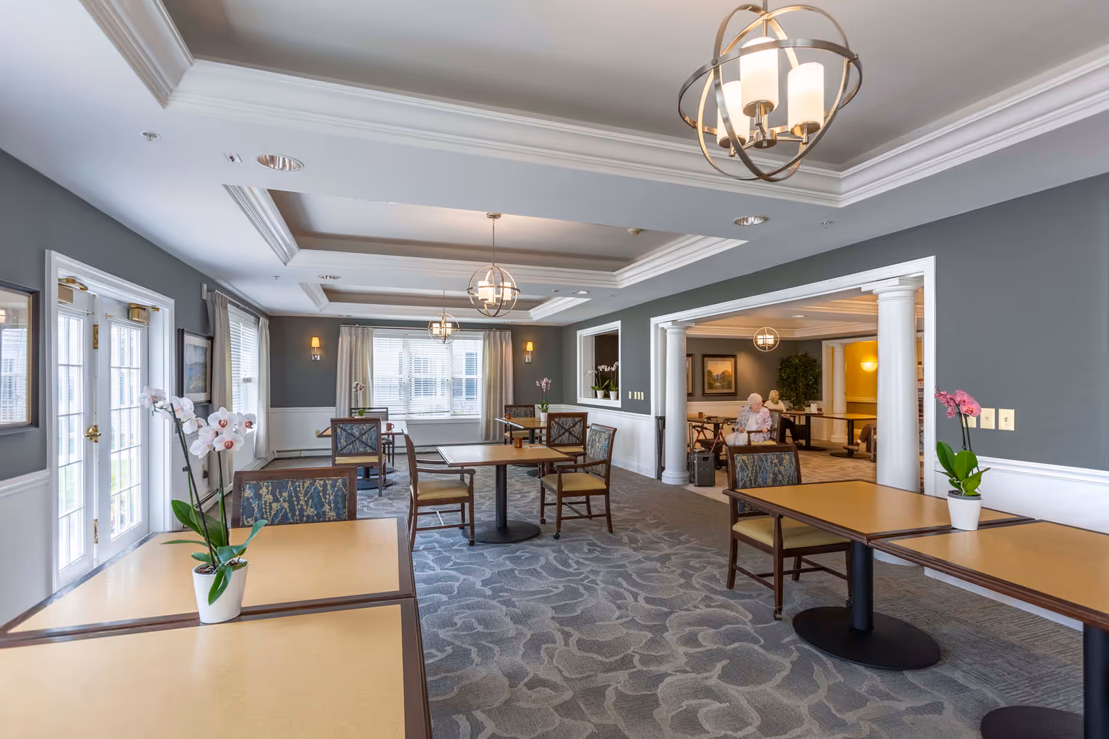 Communal dining room with tables and chairs, pendant lights, potted orchids, and two residents visible in an adjacent area.