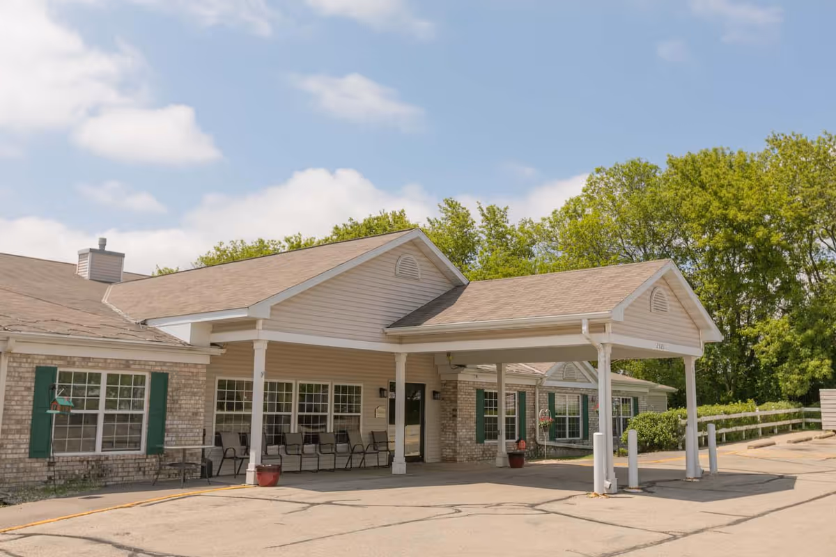 Front entrance of a single-story assisted living building with a covered porte-cochère, outdoor seating, potted plants, and trees in the background.