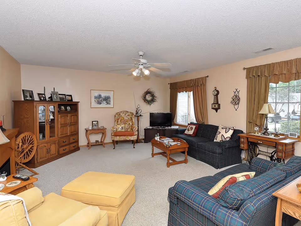 A cozy living room with beige walls and carpeted floor. The room features two plaid sofas with cushions, a yellow armchair with matching ottoman, a wooden coffee table, and a wooden cabinet with framed photos on top. There is a TV on a stand in the corner, a floral armchair, a small side table, and a vintage sewing machine table with a lamp. The windows have brown curtains, and a ceiling fan with lights is mounted on the ceiling.
