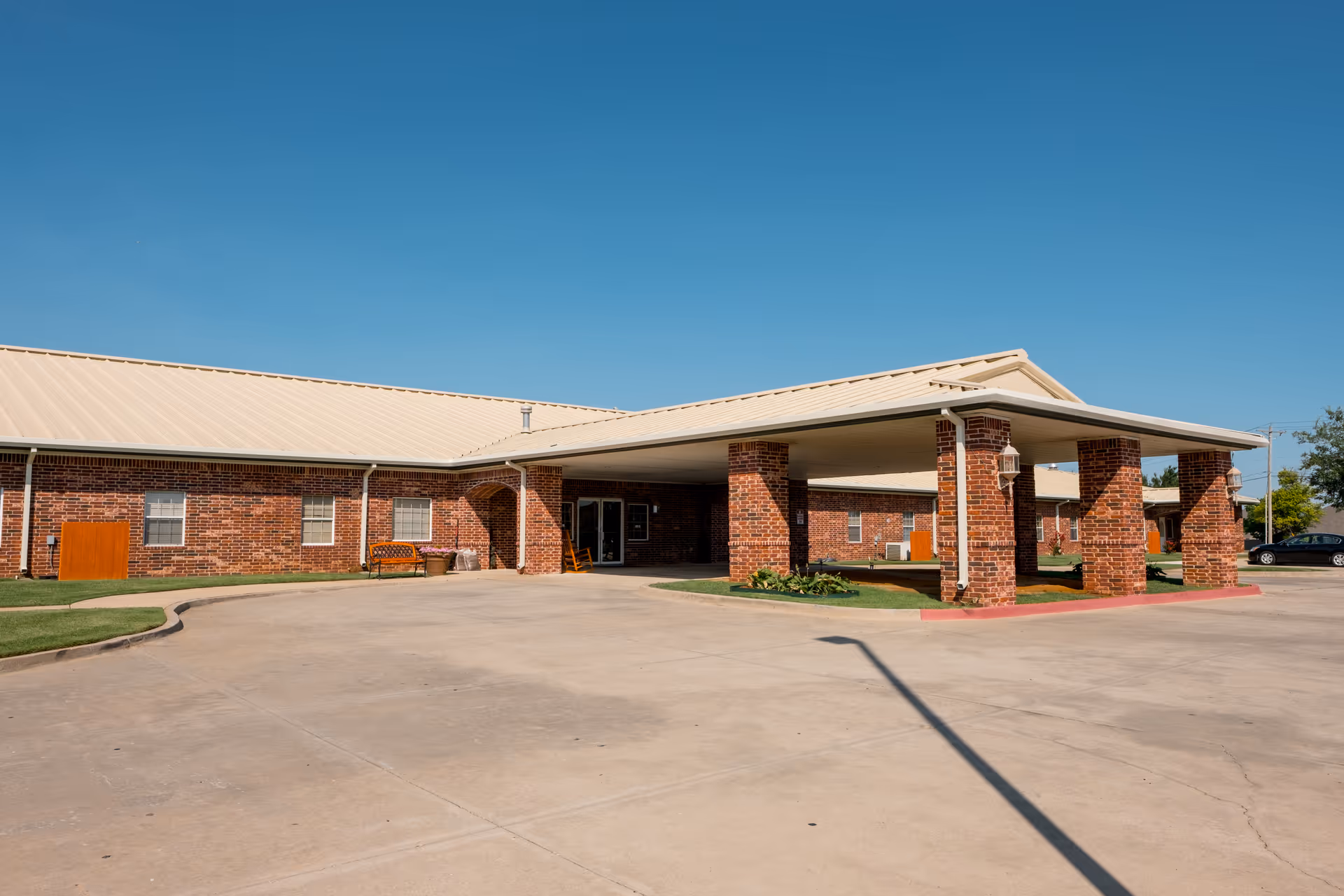 Single-story brick senior living building with a covered entrance/porte-cochère and a wide paved driveway under a clear blue sky.