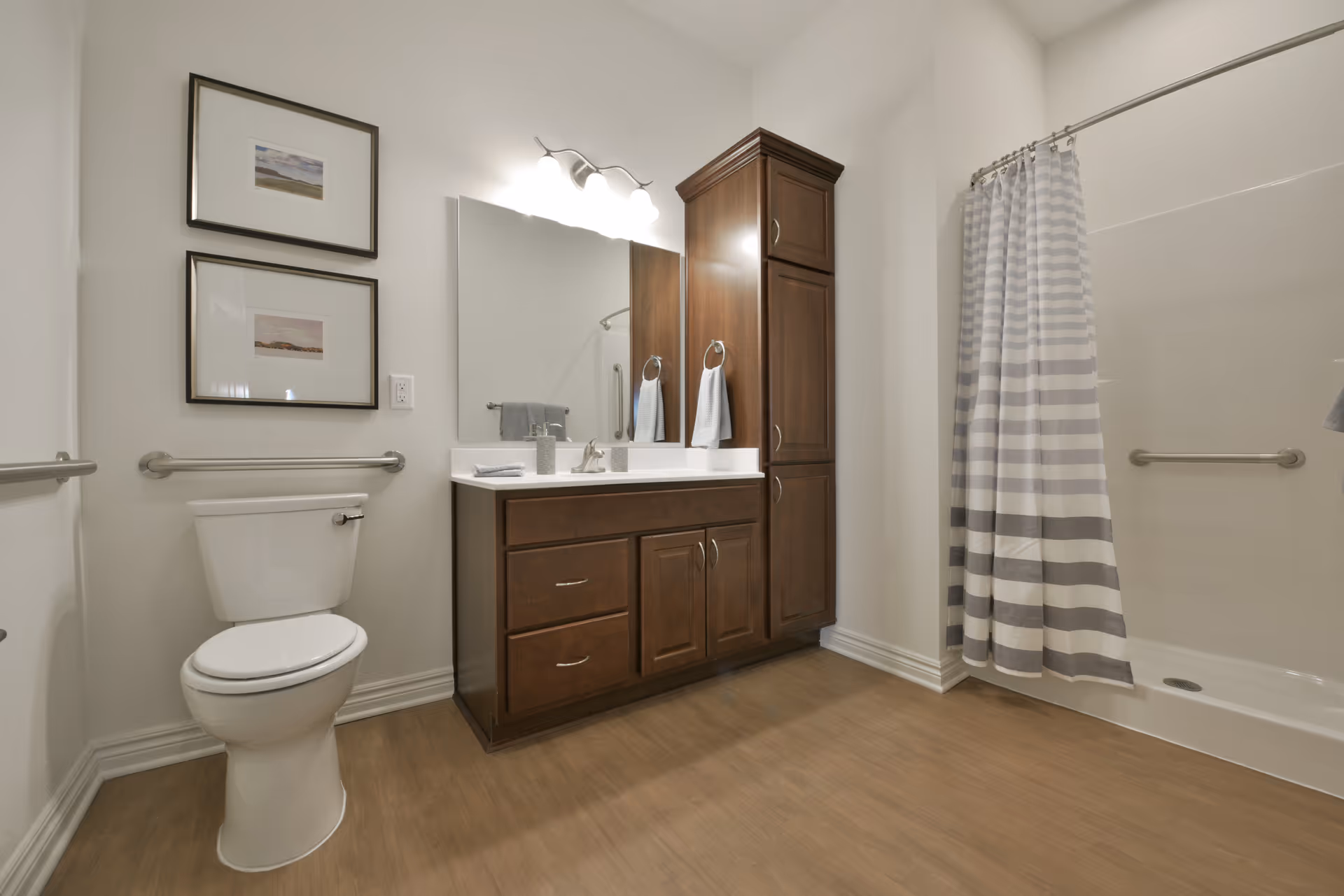 A clean and spacious bathroom featuring a white toilet with grab bars on the wall, a wooden vanity with a white countertop and sink, a large mirror above the vanity, two framed pictures on the wall, and a shower area with a striped shower curtain.