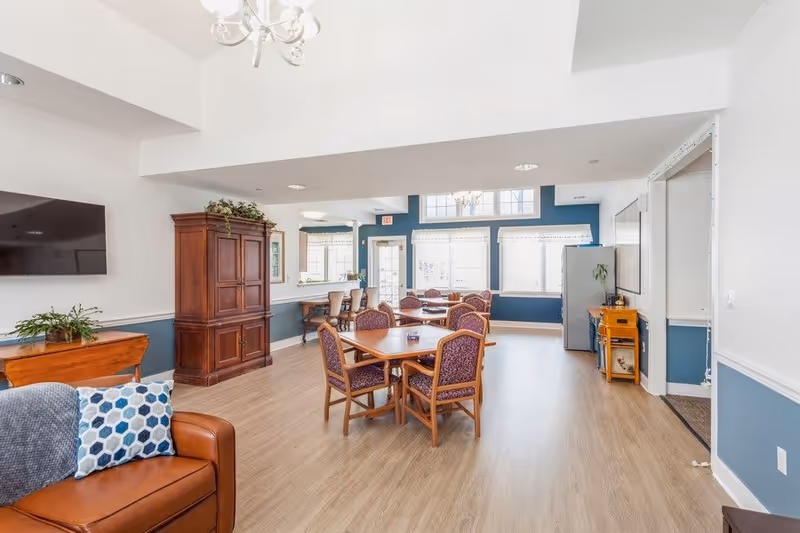 A bright and spacious common area in an assisted living facility featuring wooden tables with purple cushioned chairs, a brown leather couch with a blue and white patterned pillow, a large wooden cabinet, a flat-screen TV mounted on the wall, and large windows letting in natural light. The walls are painted white and blue, and the floor is light wood.