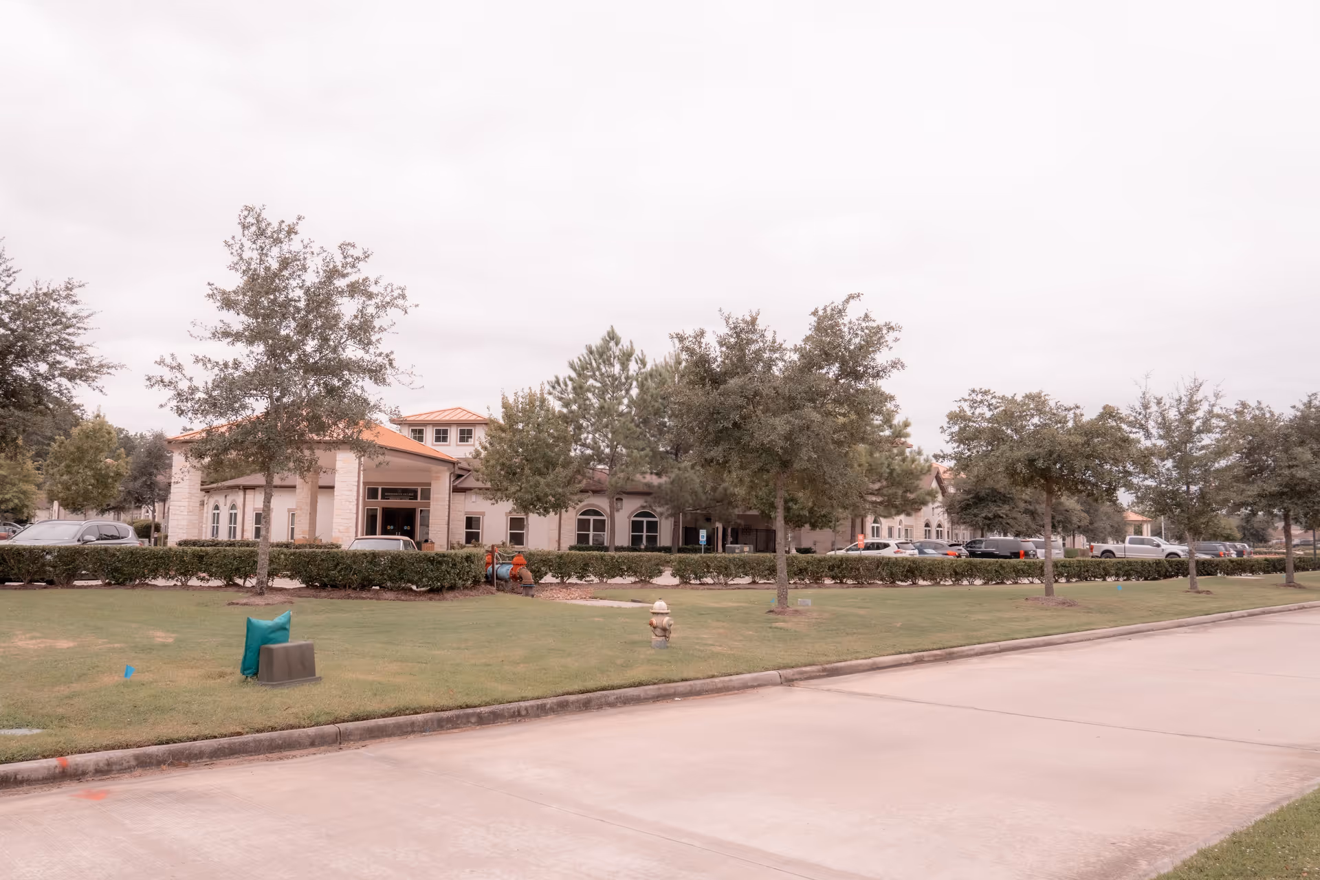 Front exterior of a senior living building with a lawn, trees, parked cars, and a driveway.