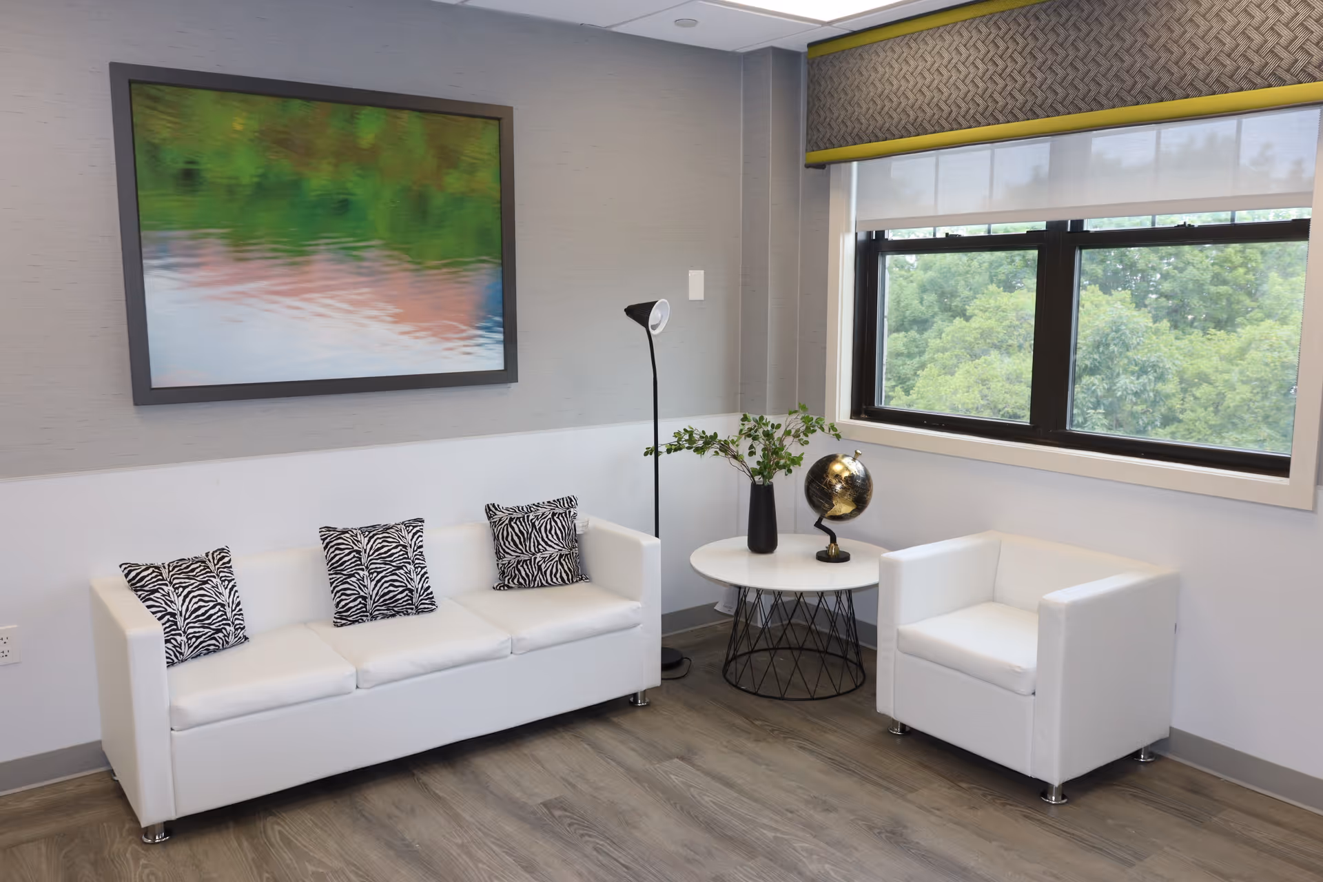 A modern sitting area with a white three-seater sofa adorned with three zebra-patterned cushions, a matching white armchair, and a round white table with a black wireframe base. On the table, there is a black vase with green foliage and a decorative globe. A tall floor lamp stands behind the table. A large window with a patterned valance shows green trees outside. A framed abstract artwork with green and pink hues hangs on the wall above the sofa.