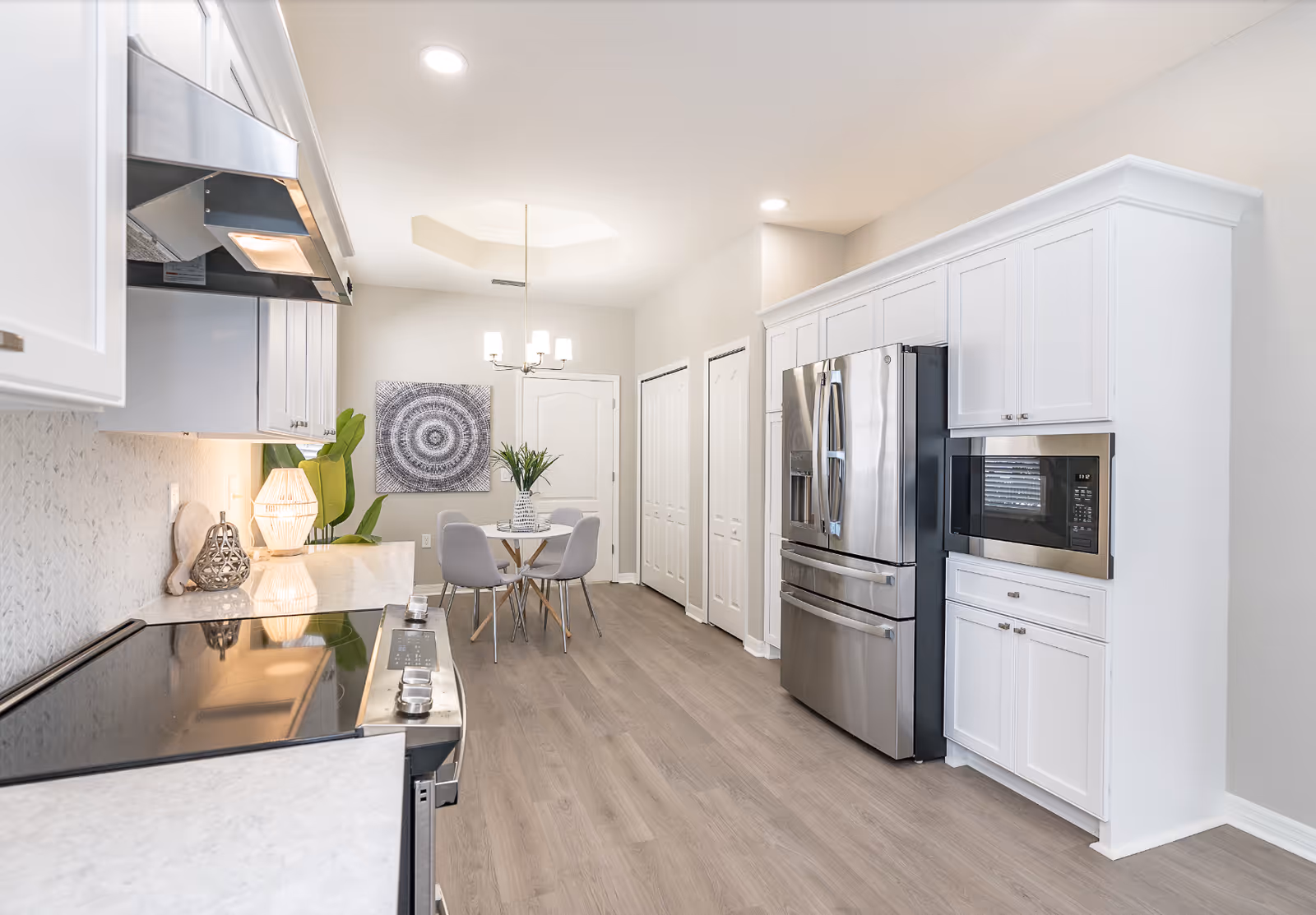 Bright modern kitchen with white cabinets, stainless steel refrigerator and microwave, a stove in the foreground, and a small dining table with chairs and artwork in the background.