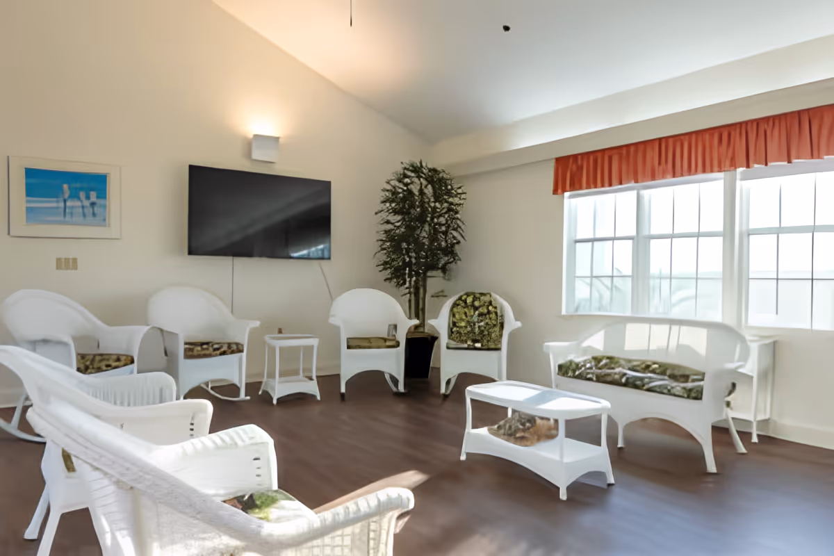 A bright living room with white wicker furniture including chairs, a loveseat, and a coffee table. The chairs have patterned cushions. There is a large flat-screen TV mounted on the wall, a potted plant in the corner, a window with an orange valance letting in natural light, and a framed picture on the wall.