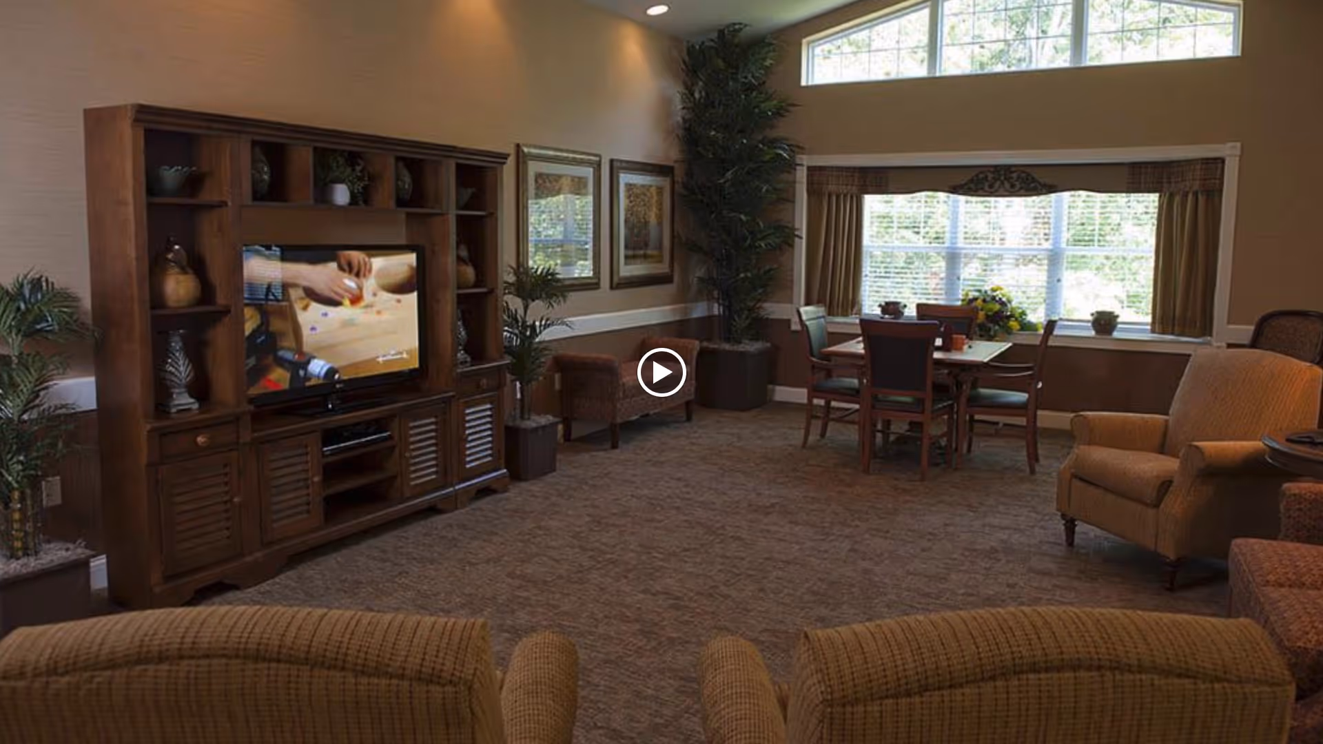 A cozy living room area with a large wooden entertainment center holding a television, two armchairs in the foreground, a dining table with chairs near a large window, and decorative plants and framed pictures on the walls.