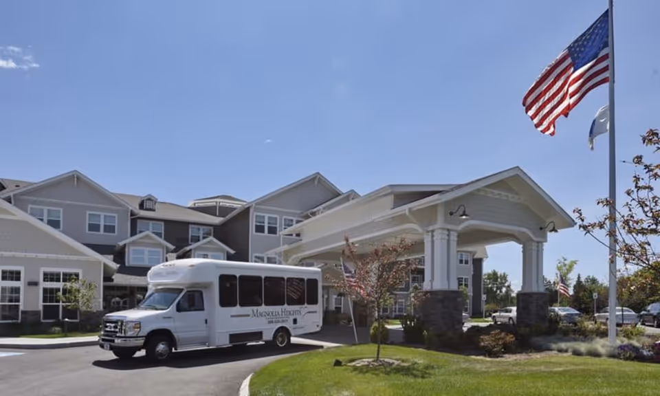 Exterior view of Magnolia Heights Gracious Retirement Living facility on a clear day, showing a large covered entrance with columns, a white shuttle bus parked in front, and an American flag flying on a flagpole.