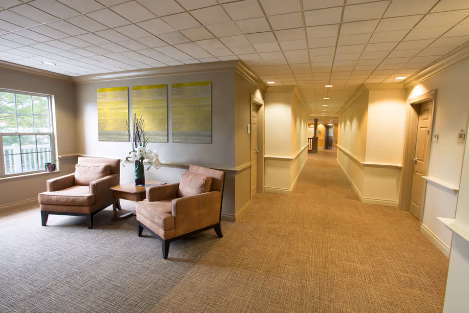 Carpeted corridor with a seating area of two armchairs and a side table with flowers beside a window and doors along the hallway.