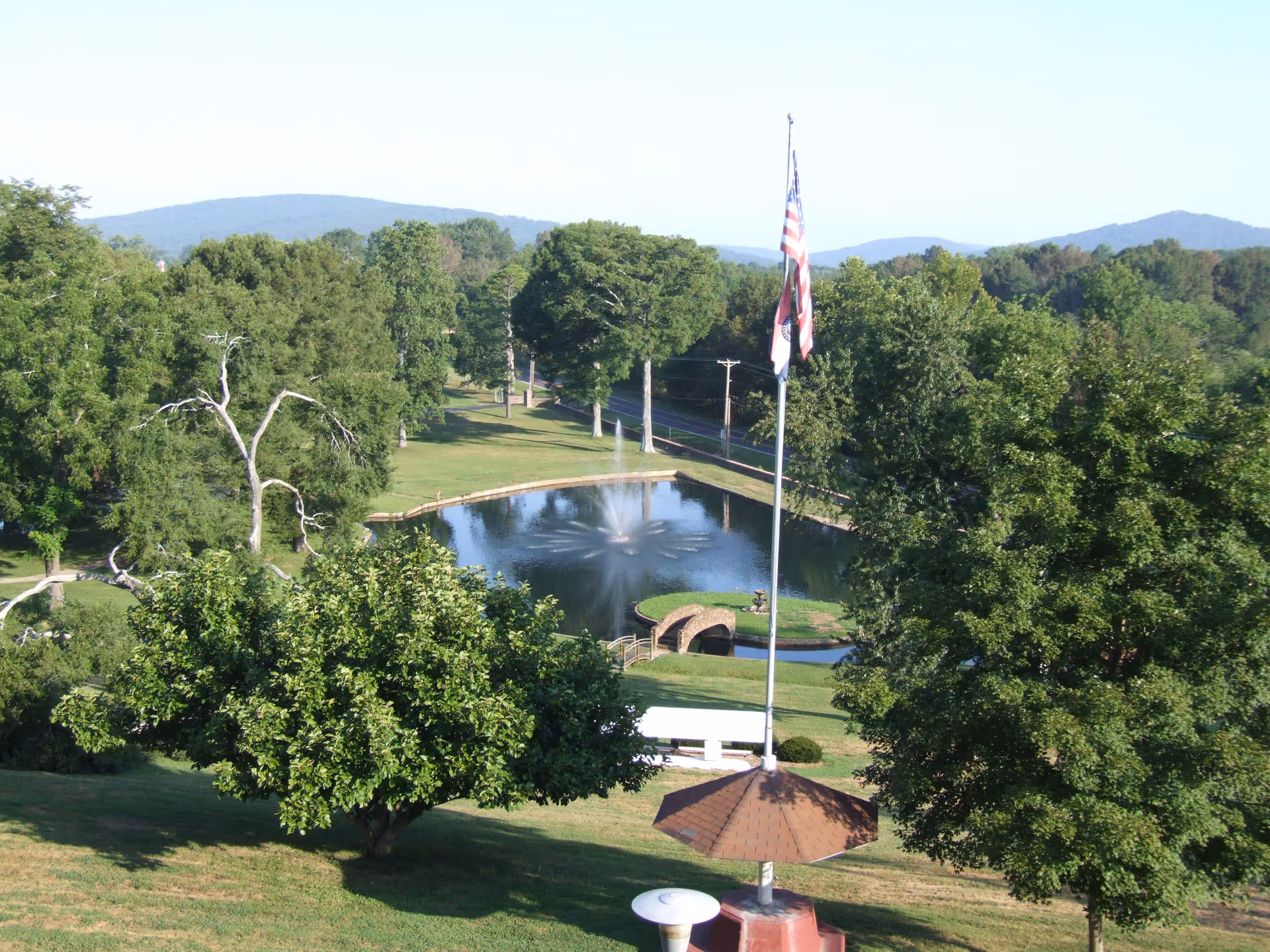 A scenic outdoor view of a park-like area with a pond featuring a water fountain in the center, surrounded by green trees and grassy hills. There is a flagpole with an American flag and another flag, a small arched bridge over the pond, and distant hills in the background under a clear sky.