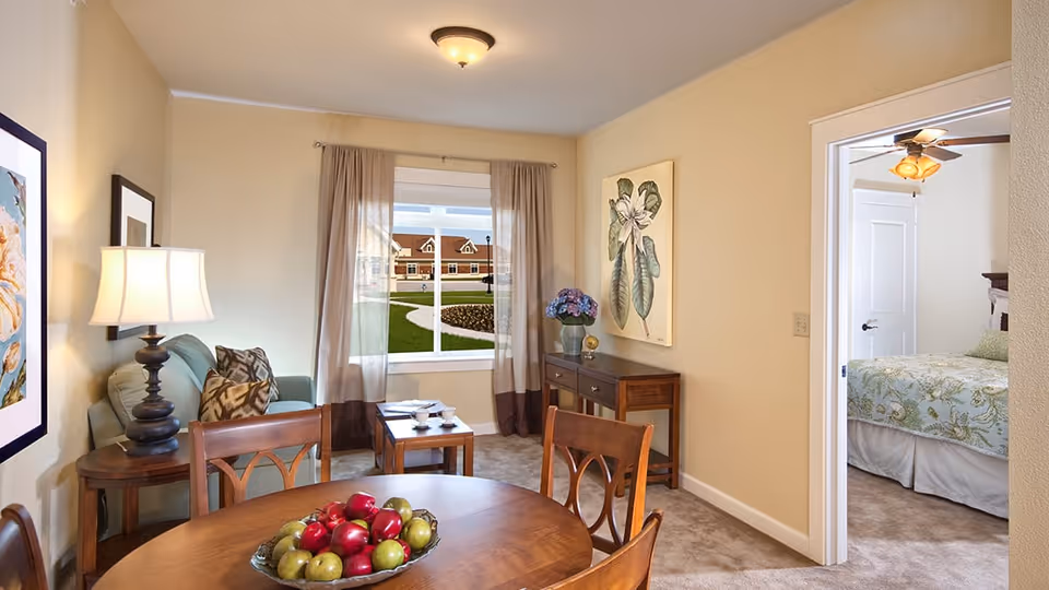 A cozy living room area with a round wooden dining table and chairs in the foreground, a small sofa with patterned pillows, a side table with a lamp, and a window with sheer curtains showing an outdoor view. A wooden console table with a vase of flowers and a large botanical painting is against the wall. Through an open doorway, a bedroom with a bed, ceiling fan, and white door is visible.