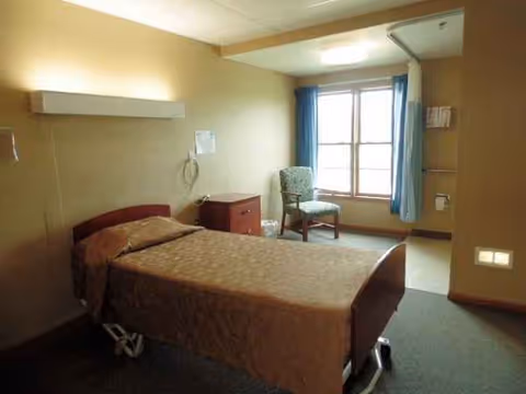 A hospital-style bedroom in a senior living facility with bed, chair, and window.