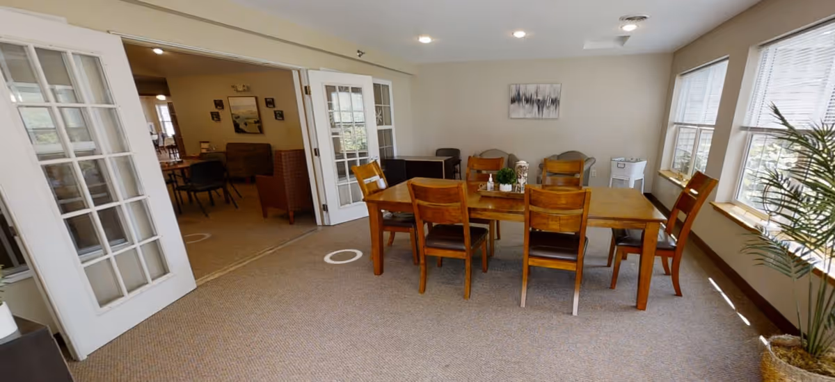 A bright dining area in a senior living facility with a wooden table and six chairs. The room has large windows with blinds allowing natural light to fill the space. There are two armchairs and a small table against the far wall, a potted plant near the window, and double glass doors leading to another room with additional seating.