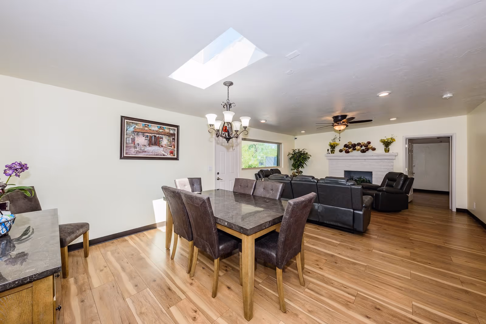 A bright and spacious living and dining area with wooden flooring. The dining area features a rectangular table with six dark cushioned chairs under a chandelier. The living area has black leather sofas arranged around a white brick fireplace decorated with flowers and wall art. A ceiling fan is mounted above the living area, and a skylight provides natural light.