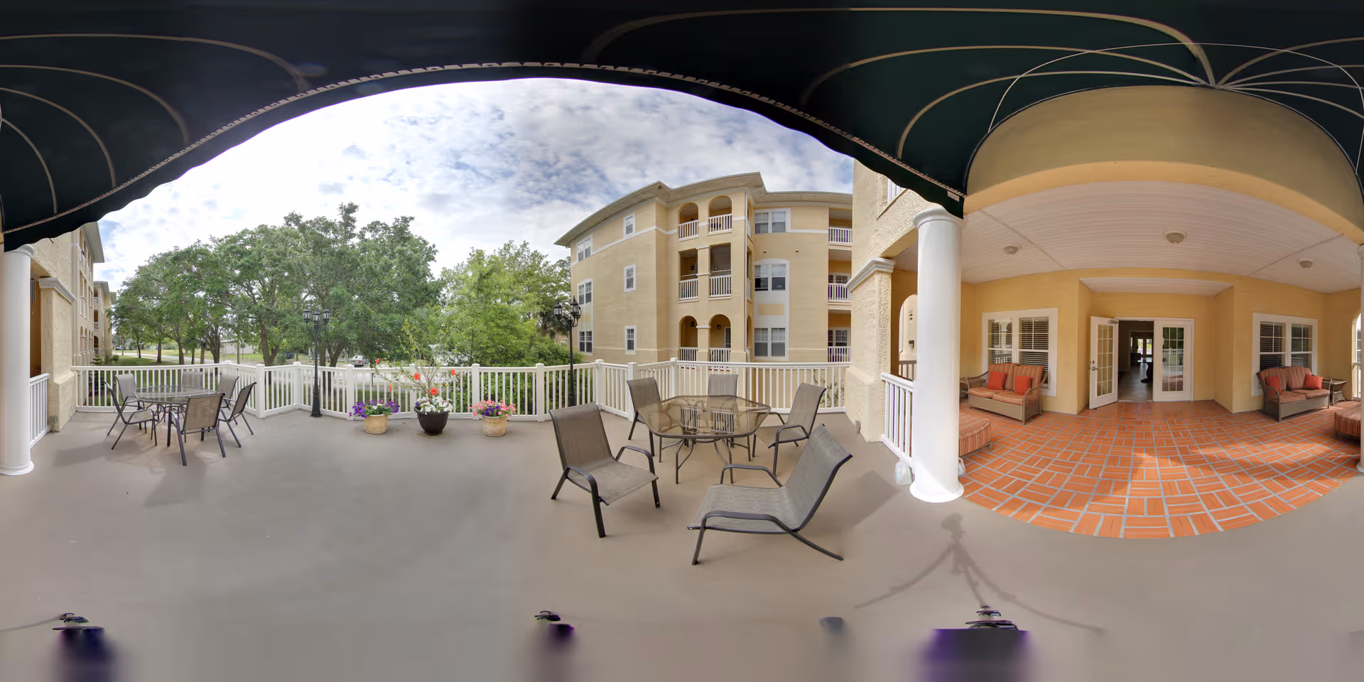 Outdoor patio area at a senior living facility with several tables and chairs arranged for seating. The patio is covered with a dark green awning and has a white railing. There are potted plants with flowers along the railing. In the background, there are trees and a multi-story beige building with balconies. The patio also has a tiled section with cushioned seating near the building entrance.