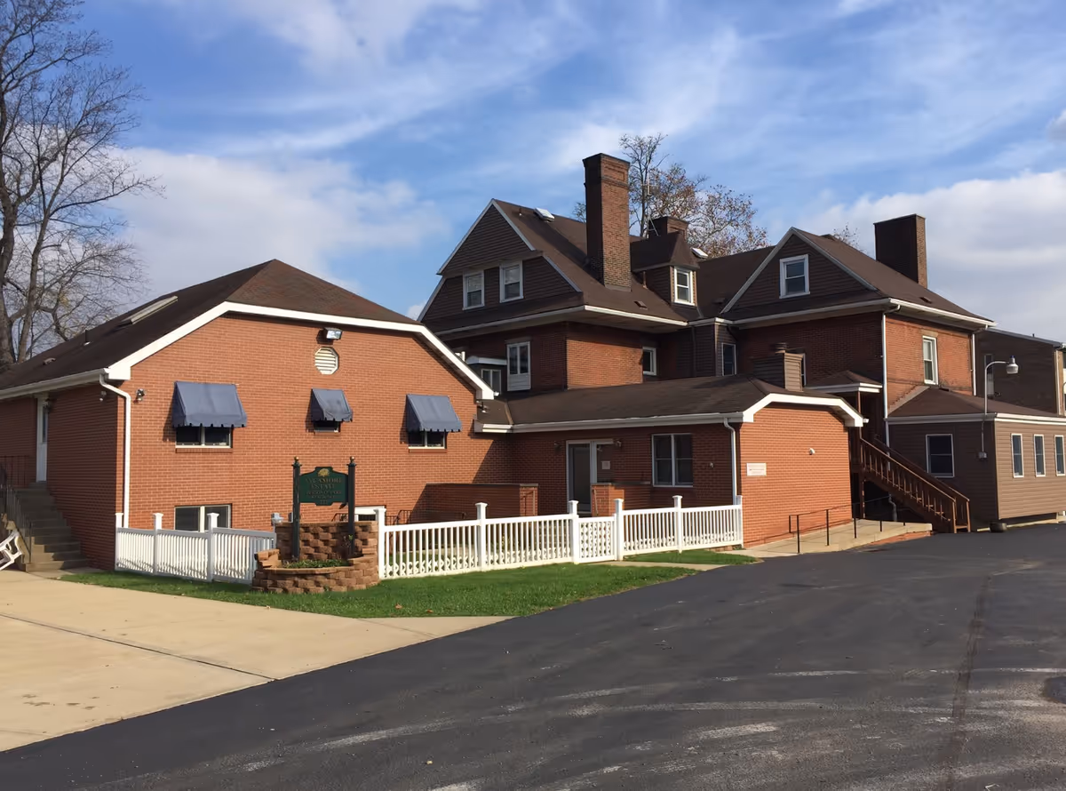 Exterior view of Sycamore Estate Inc., a multi-story brick building with brown roofing and several chimneys. The building has blue awnings over some windows and a white fence enclosing a small grassy area with a sign in front. The sky is partly cloudy.