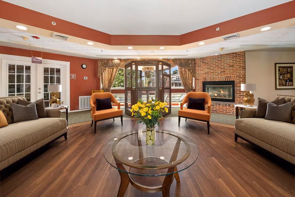 A cozy living room area with two beige sofas and two orange armchairs arranged around a round glass coffee table with a vase of yellow flowers. The room features wood flooring, a brick fireplace on the right wall, large windows with drapes, and a door leading outside. The walls are painted in warm tones with recessed ceiling lights providing illumination.