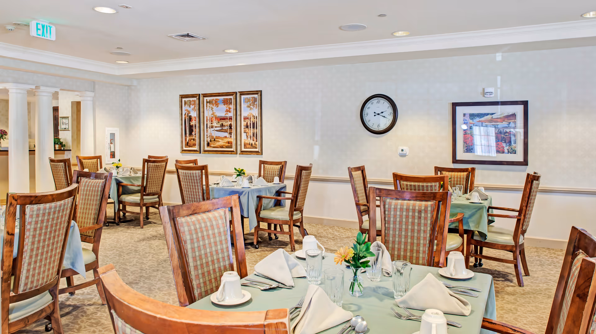 Bright dining room with multiple round tables set with napkins, glassware, and wooden chairs.