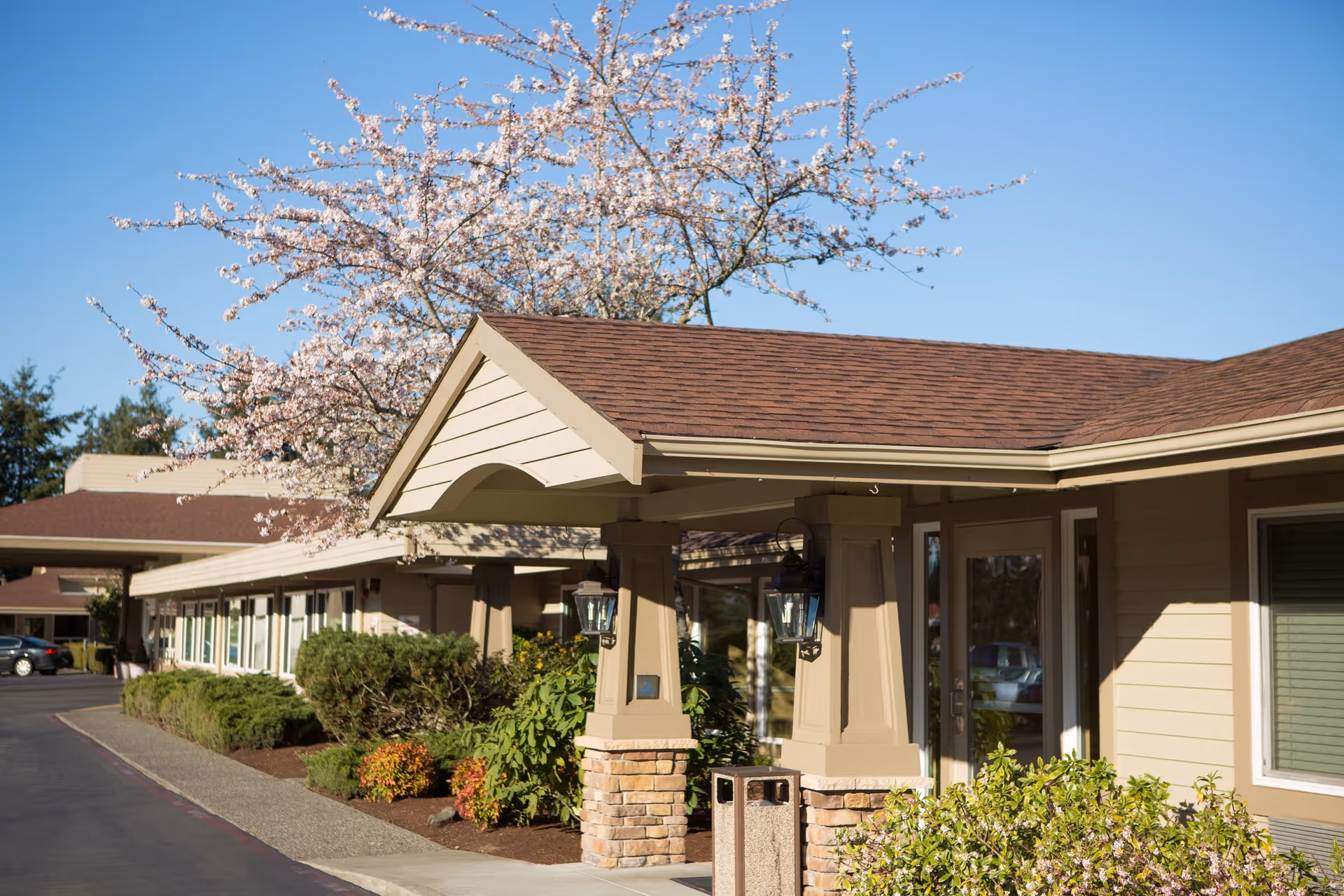 Front exterior and covered entrance of a single-story senior living building with landscaping and a blooming tree.