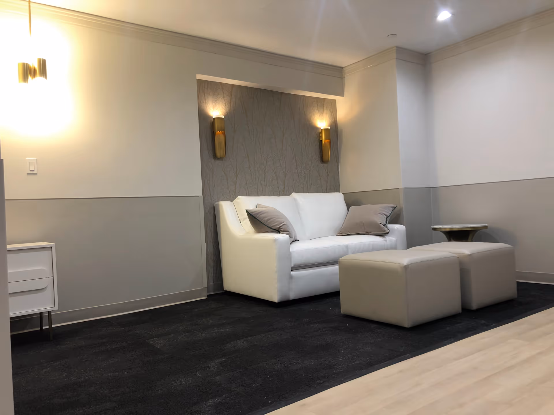 A modern sitting area with a white couch adorned with two gray pillows, two beige ottomans, a small round side table, and a white cabinet against a wall with two gold wall sconces providing warm lighting.