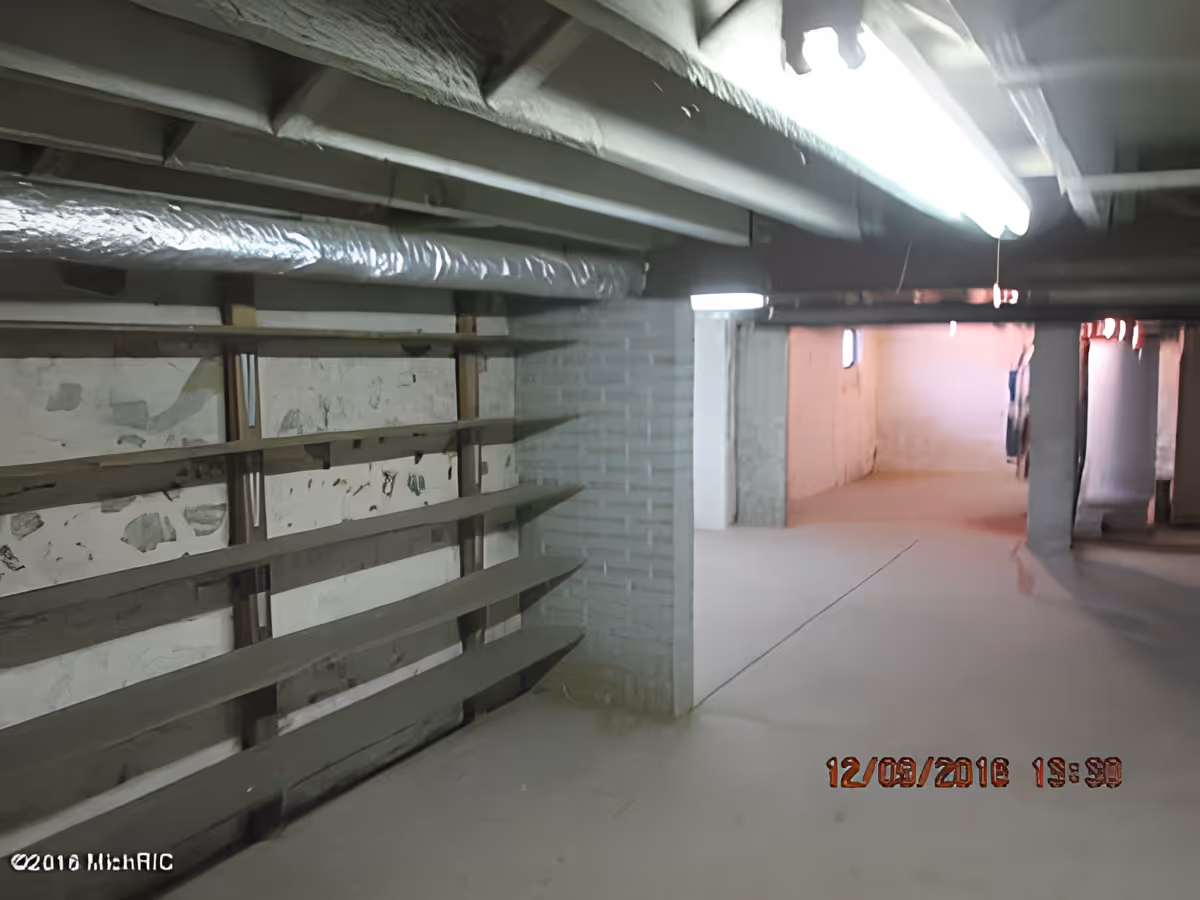Empty basement-like interior with exposed ceiling beams, support columns, concrete floor and wooden shelving.