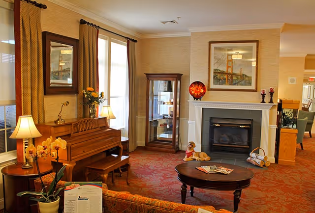 A cozy living room with a wooden piano against the wall, a small wooden bench, a round wooden coffee table with magazines, a fireplace with decorative items on the mantel, a framed picture above the fireplace, a glass display cabinet, and large windows with curtains letting in natural light. There are also lamps, flowers, and comfortable seating visible.