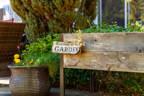 A raised wooden garden bed filled with green plants and a small wooden sign that reads 'Welcome to our GARDEN'. Next to it is a large brown planter with yellow and pink flowers. In the background, there are bushes and a tree, with a building window partially visible.