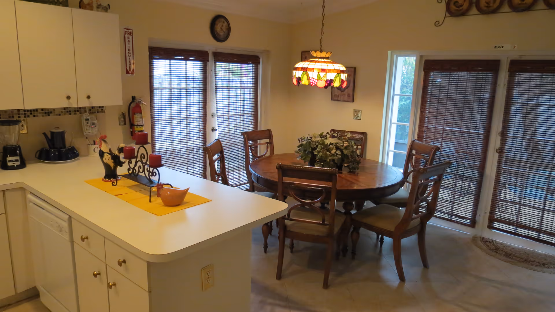 Interior view of a dining area adjacent to a kitchen. The dining area features a round wooden table with six chairs and a decorative hanging lamp above it. The kitchen counter has a white surface with decorative items including a candle holder and a small ceramic bowl. There are two sets of glass doors with bamboo blinds allowing natural light into the room.