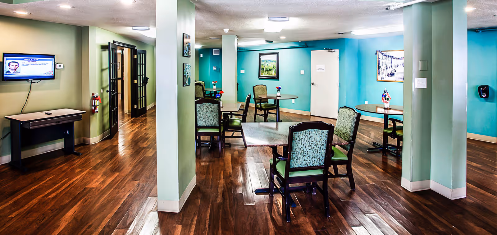 Interior view of a senior living facility common area with wooden floors, green and teal walls, several round tables with chairs, small flower vases on the tables, a wall-mounted TV, and framed artwork on the walls.