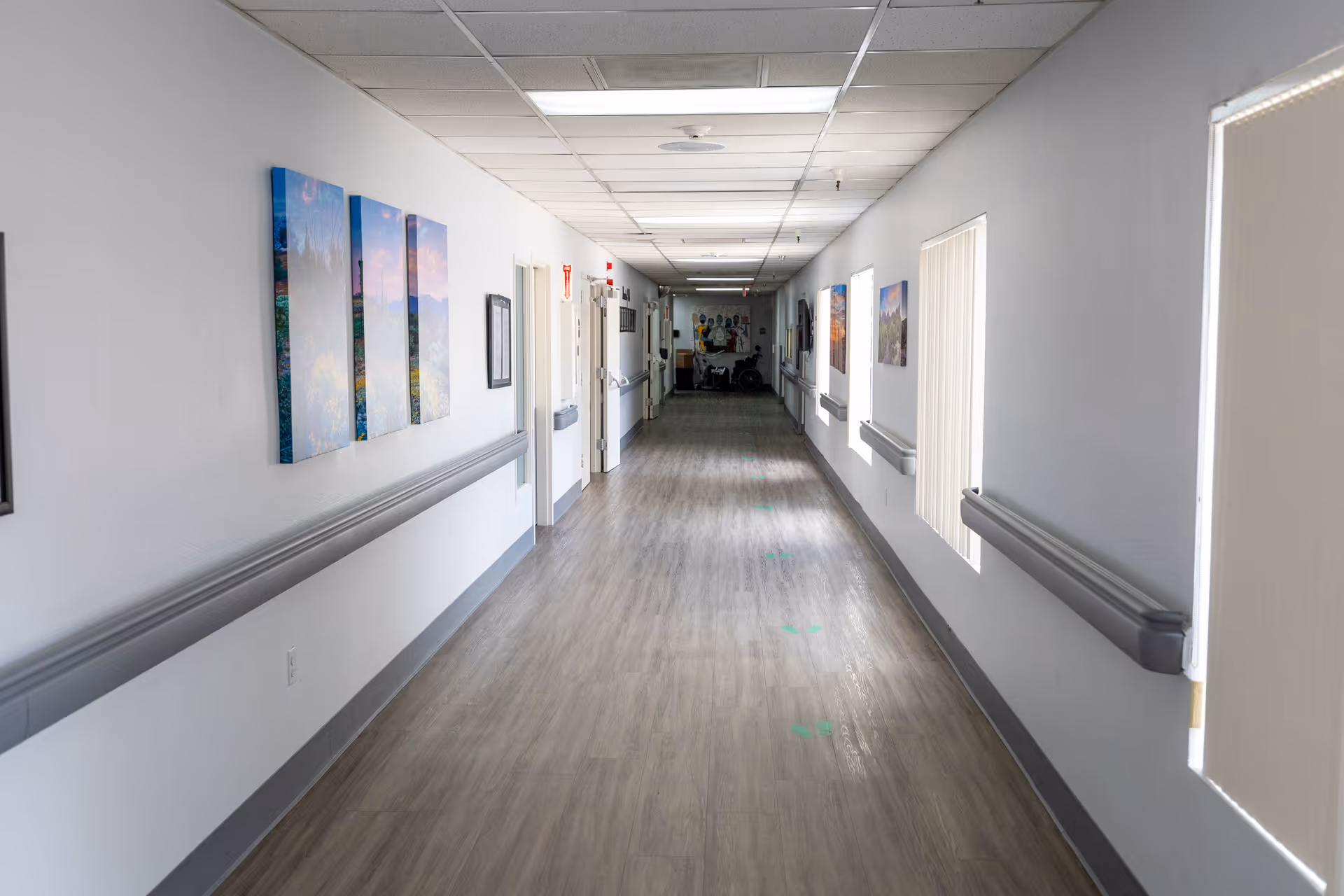 A long, clean hallway in a senior living facility with light gray walls, handrails on both sides, and several doors along the corridor. The floor is wood-patterned, and there are framed pictures and windows with blinds on the right wall. The ceiling has white tiles with fluorescent lighting.