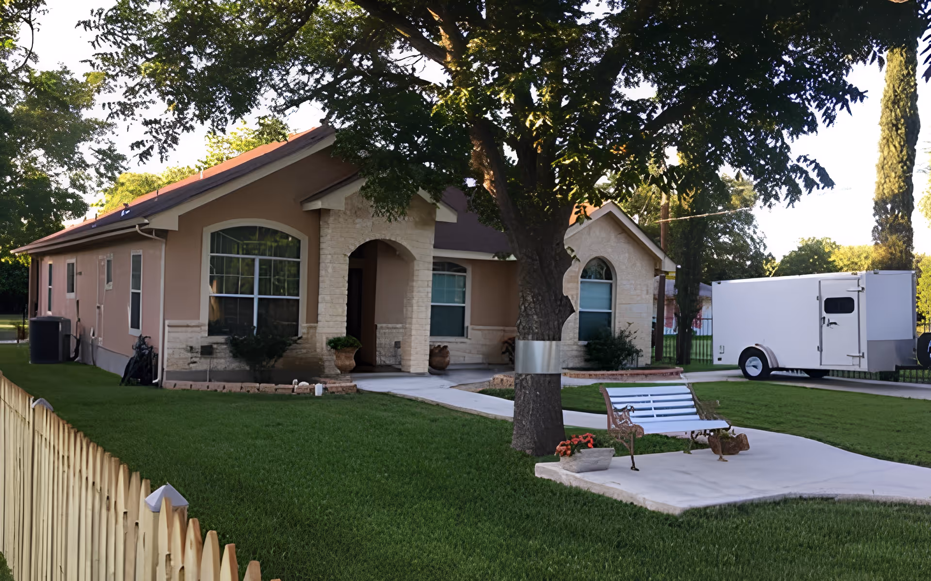 Exterior view of a single-story residential building with beige walls and a brown roof, surrounded by green grass and trees. A white bench and flower pots are placed on a concrete path in the front yard. A white trailer is parked on the driveway to the right of the building.
