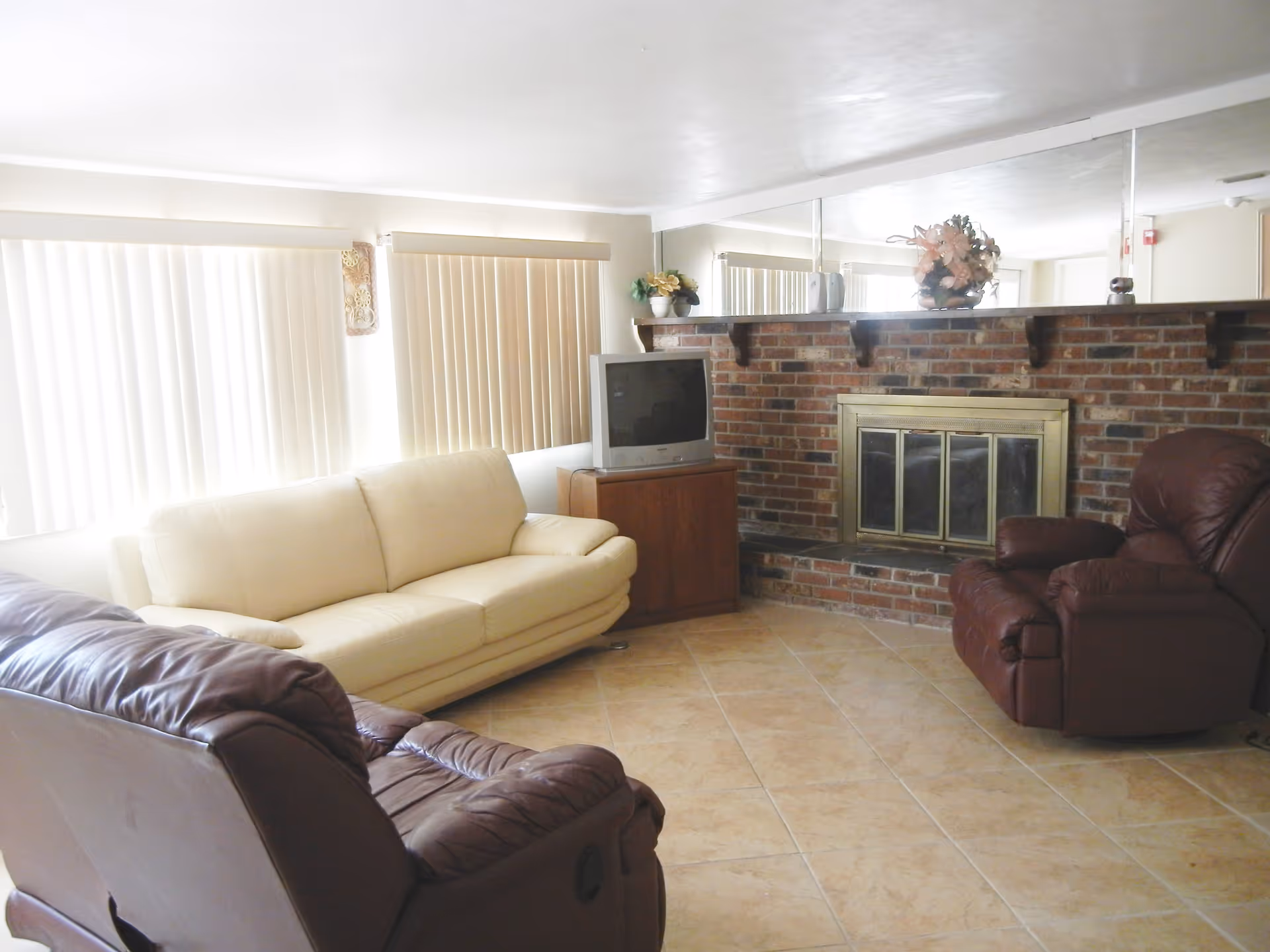 A living room with a cream-colored sofa, two brown leather recliners, a brick fireplace with a brass-framed glass door, a small wooden TV stand with an older model television, and a large mirror above the fireplace. The room has tiled flooring and vertical blinds covering the windows.