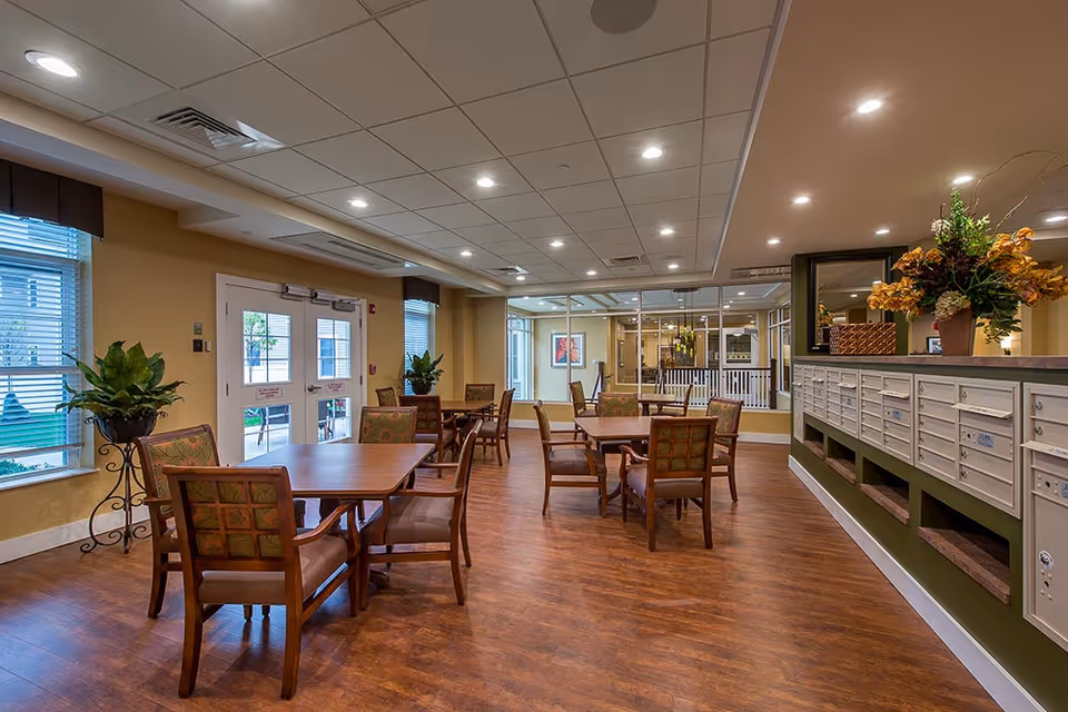 A well-lit interior room with several wooden tables and chairs arranged for seating. The room has large windows with blinds, potted plants, and a row of mailboxes along one wall topped with a decorative flower arrangement. The floor is wooden, and the ceiling has recessed lighting.