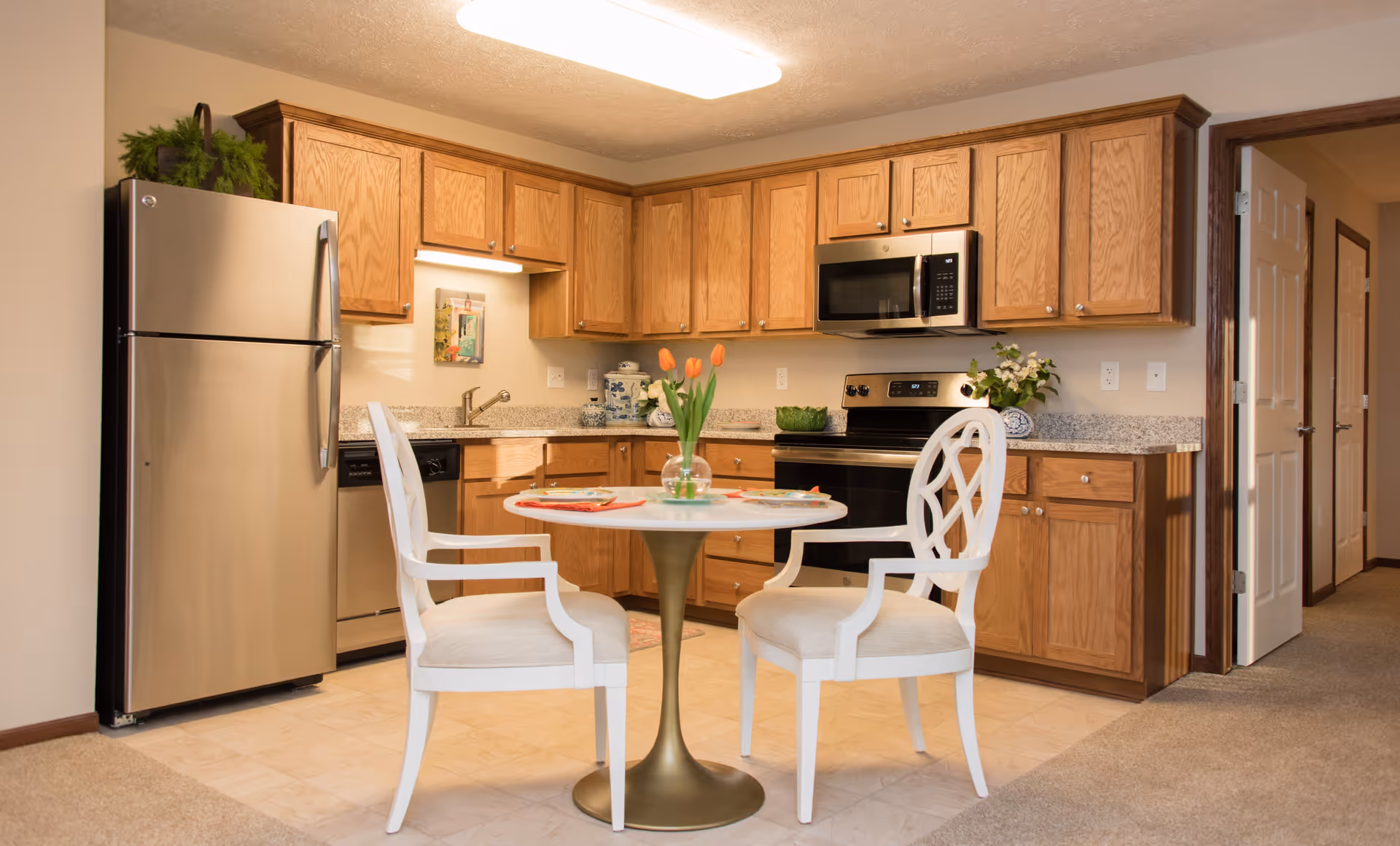 A cozy kitchen area with wooden cabinets, a stainless steel refrigerator, dishwasher, stove, and microwave. In the center is a small round table with a gold base, set with two white cushioned chairs and a vase of orange tulips. The floor is a combination of tile and carpet, and there is a hallway with open doors visible to the right.