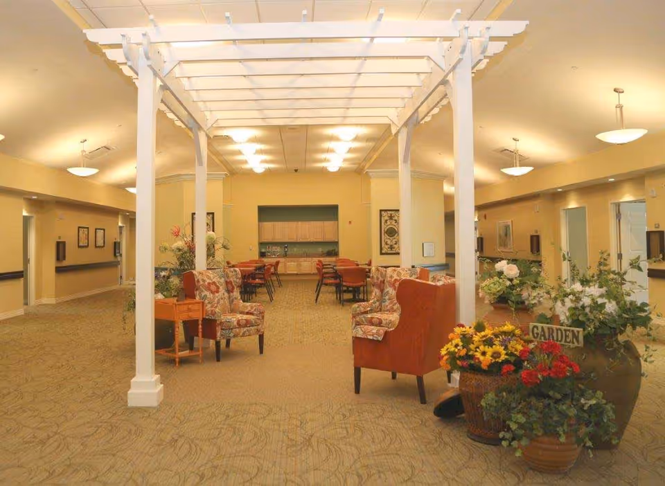 A spacious, well-lit common area in a senior living facility with floral upholstered armchairs, a white pergola structure, several tables and chairs in the background, and large pots of colorful flowers labeled 'GARDEN'. The walls are painted a soft yellow and the ceiling has recessed lighting.