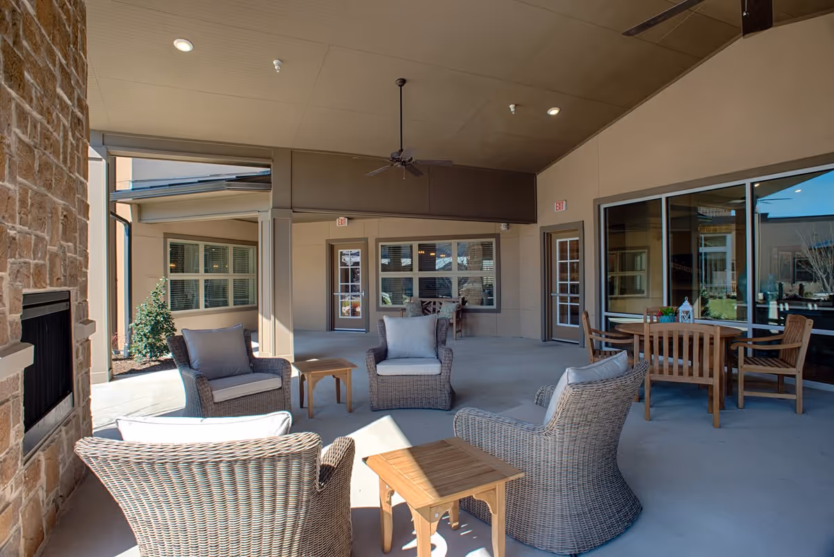 Covered outdoor seating area with wicker chairs and small wooden tables arranged around a stone fireplace. There is a ceiling fan above and a wooden dining table with chairs near large windows. The area is part of a senior living facility with beige walls and multiple doors leading inside.