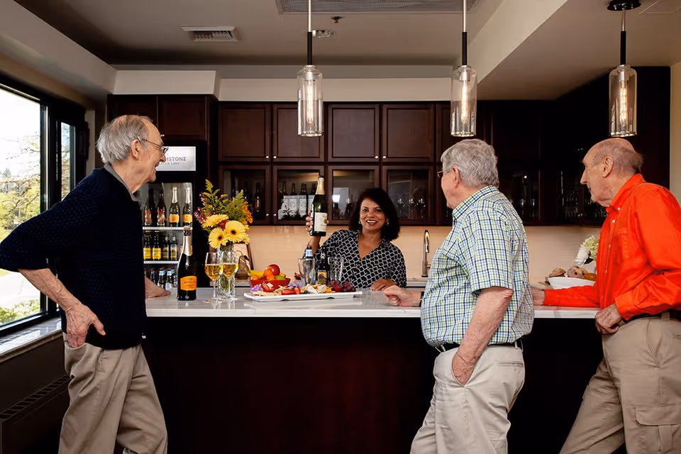 A woman behind a kitchen counter smiling and holding up a bottle of wine while three elderly men stand on the other side of the counter engaging with her. The kitchen has dark wooden cabinets, pendant lights, and a window letting in natural light. There are flowers, wine glasses, and a tray with fruit on the counter.