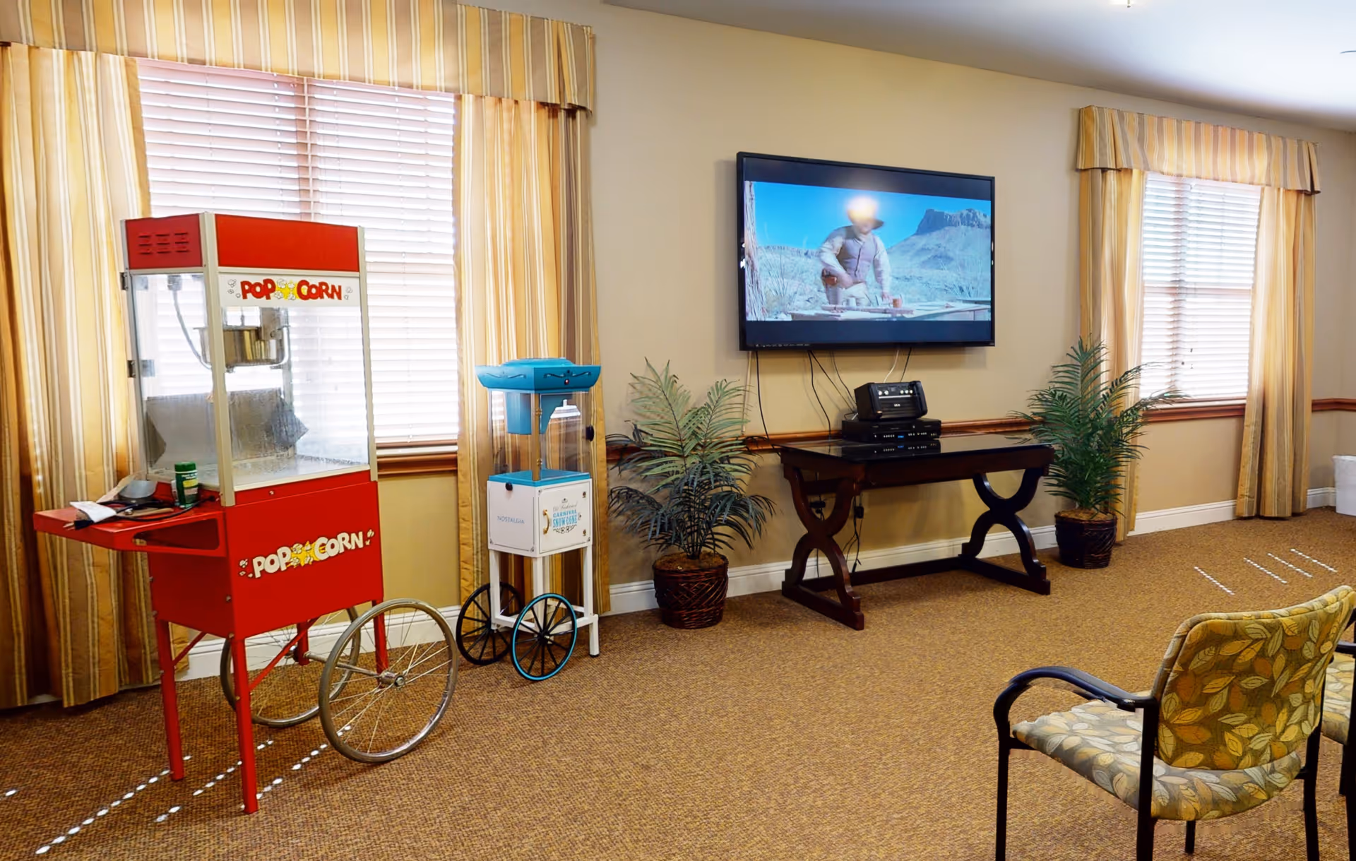 A cozy common area with a red popcorn machine on wheels, a blue and white water dispenser, two potted plants, a wall-mounted flat screen TV showing a western scene, and a wooden table holding electronic devices. The room has beige walls, striped curtains on the windows, and patterned chairs.