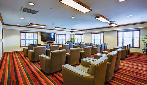 A bright lounge/media room with rows of beige armchairs facing a wall-mounted TV and large windows.