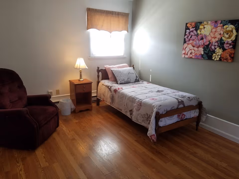 A small bedroom with a single wooden bed covered with a Paris-themed bedspread and pillows. Next to the bed is a wooden nightstand with a lamp and a small trash bin on the floor. A maroon upholstered armchair is positioned to the left. The room has hardwood floors, a window with a brown valance, and a colorful floral painting on the wall.