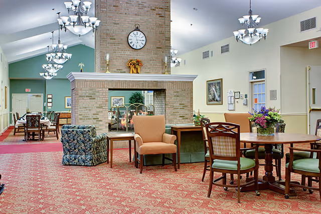 A spacious senior living common area with a large brick double-sided fireplace in the center. The room features multiple seating arrangements including cushioned armchairs and wooden chairs around tables. The floor is covered with a red patterned carpet, and the walls are painted in soft green and beige tones. Several chandeliers hang from the ceiling, and there are framed pictures and a clock on the fireplace. A vase with flowers is placed on one of the tables.