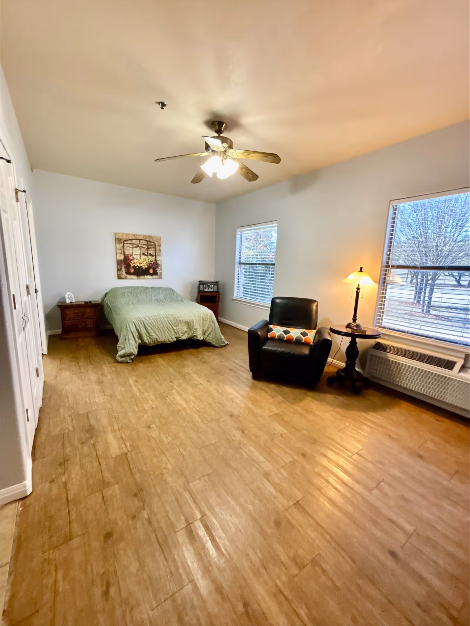 A bedroom with light wooden flooring, a bed with a green bedspread against the wall, a wooden nightstand with a clock, a black armchair with a colorful pillow, a round side table with a lamp, two windows with white blinds, and a ceiling fan with lights.