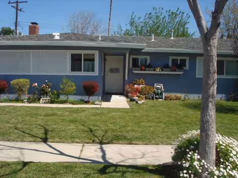 A single-story blue senior living facility exterior with a front lawn and flower beds.