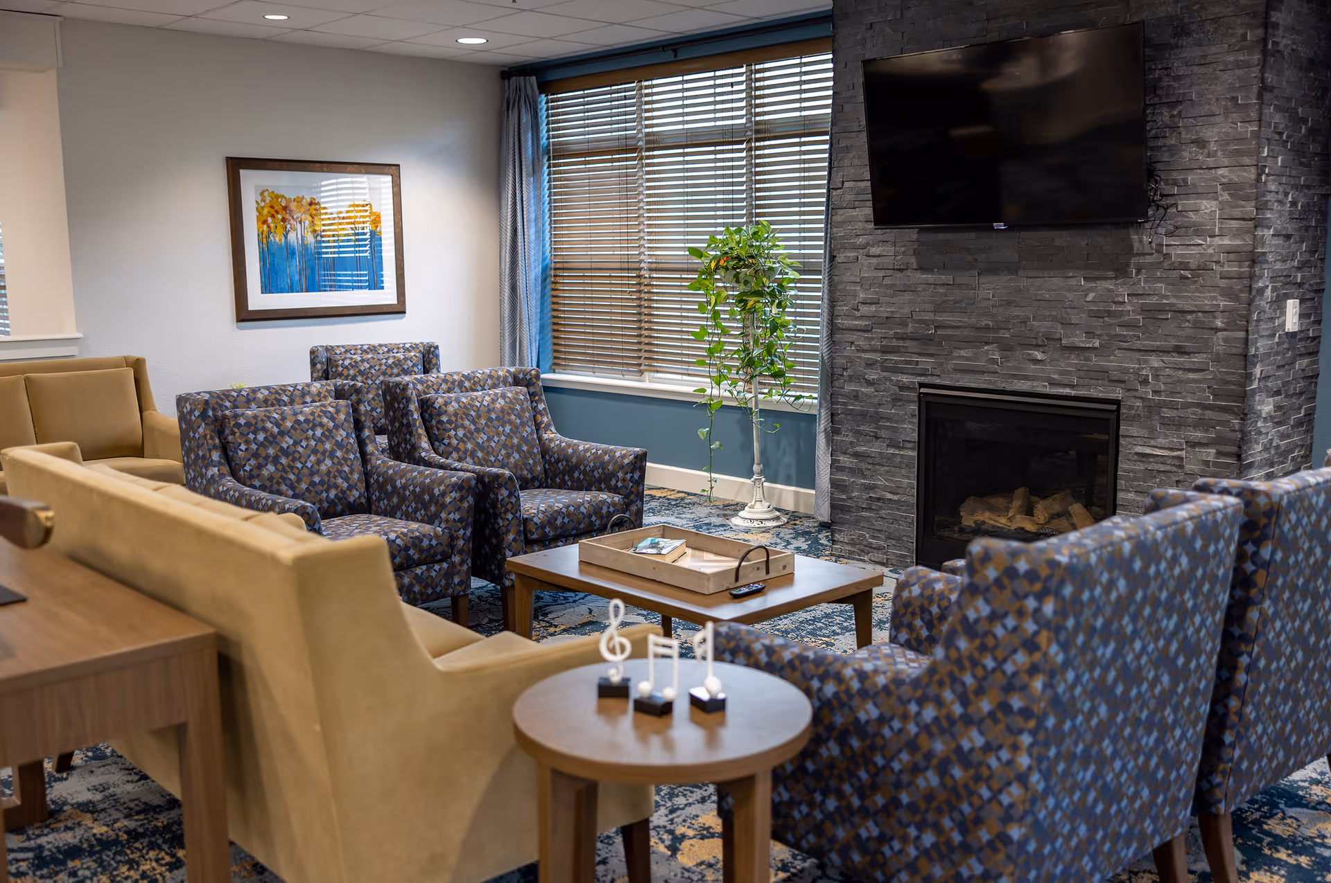A cozy living room area with patterned armchairs and beige sofas arranged around a wooden coffee table. A flat-screen TV is mounted above a modern stone fireplace. A window with blinds and curtains lets in natural light, and a potted plant stands beside the window. A small round table in the foreground holds decorative musical note sculptures.