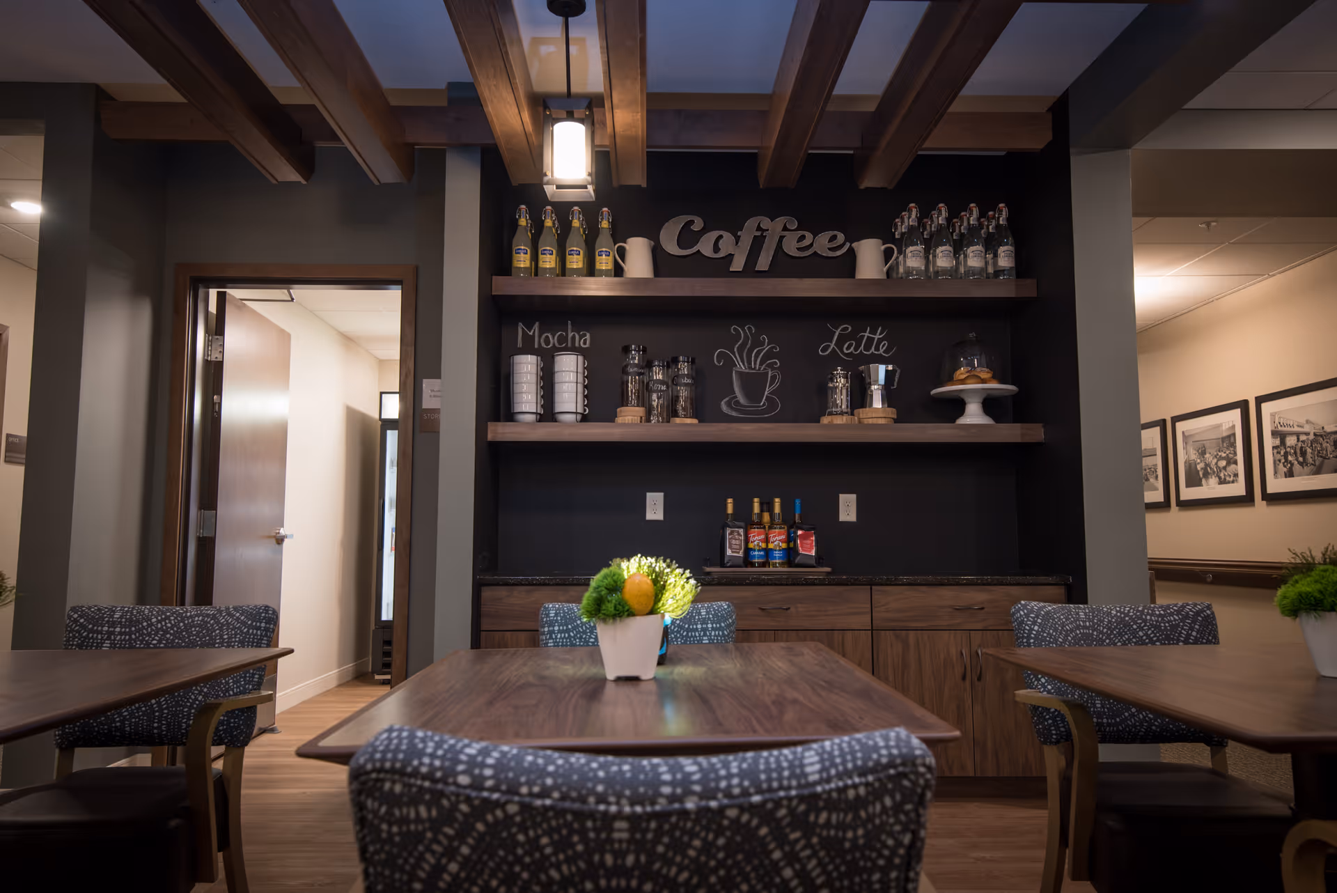 Interior view of a coffee station area in a senior living facility with wooden tables and patterned chairs. The coffee station has shelves with bottles, mugs, and jars labeled Mocha and Latte, with a decorative 'Coffee' sign above. There are small potted plants on the tables and framed pictures on the wall in the hallway.