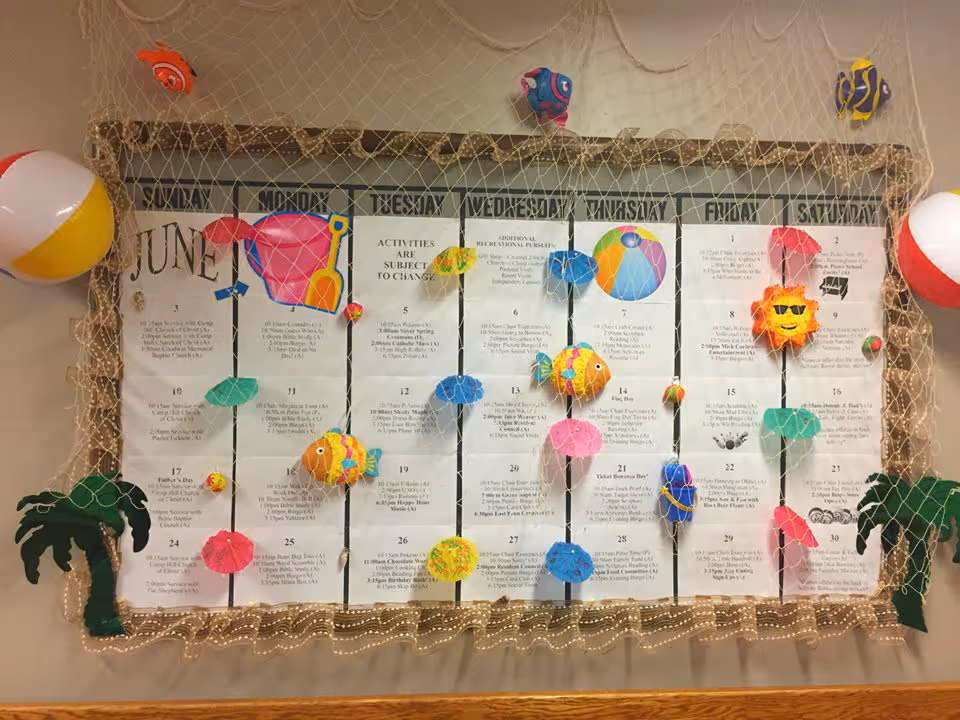 A large June calendar displayed on a wall, decorated with a fishing net, colorful paper fish, beach balls, umbrellas, a sun with sunglasses, and palm trees, creating a summer beach theme.