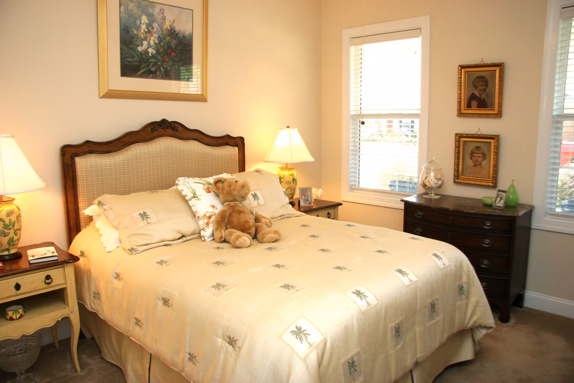 A cozy bedroom with a large bed covered in a beige bedspread featuring palm tree patterns. A teddy bear sits on the bed. There are two bedside tables with lamps on either side of the bed. The room has two windows with white blinds, a wooden dresser with framed portraits above it, and a framed floral painting hanging on the wall above the bed.