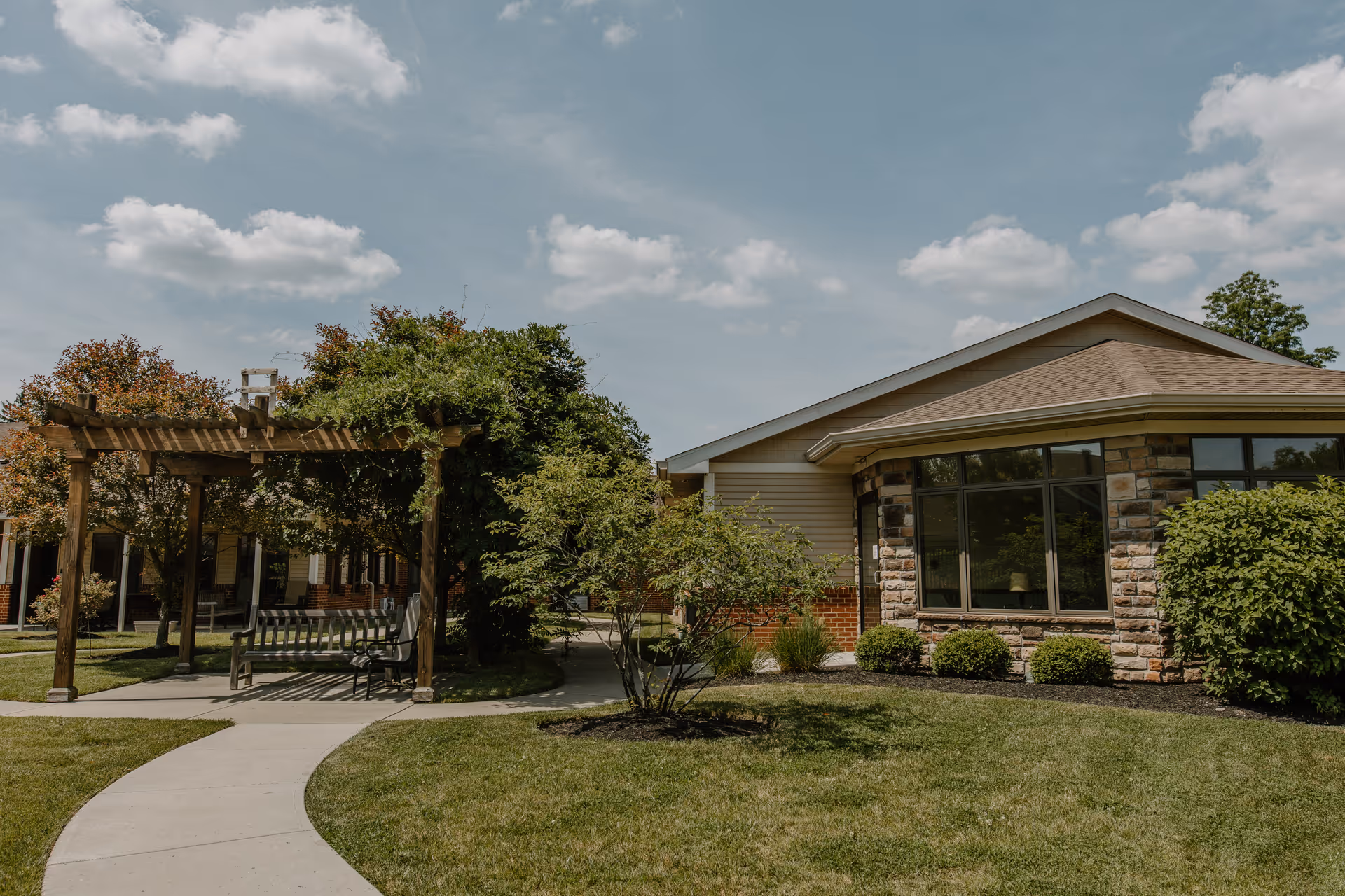 Outdoor view of a senior living facility with a curved concrete pathway leading to a wooden pergola with benches underneath. The building has a stone and siding exterior with large windows, surrounded by green grass, bushes, and trees under a partly cloudy sky.
