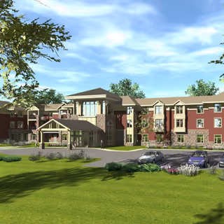Exterior view of a large senior living facility named Fieldstone at Chester Springs, featuring a three-story building with a covered entrance, stone and brick facade, surrounded by green lawns, trees, and a parking area with several cars under a partly cloudy sky.