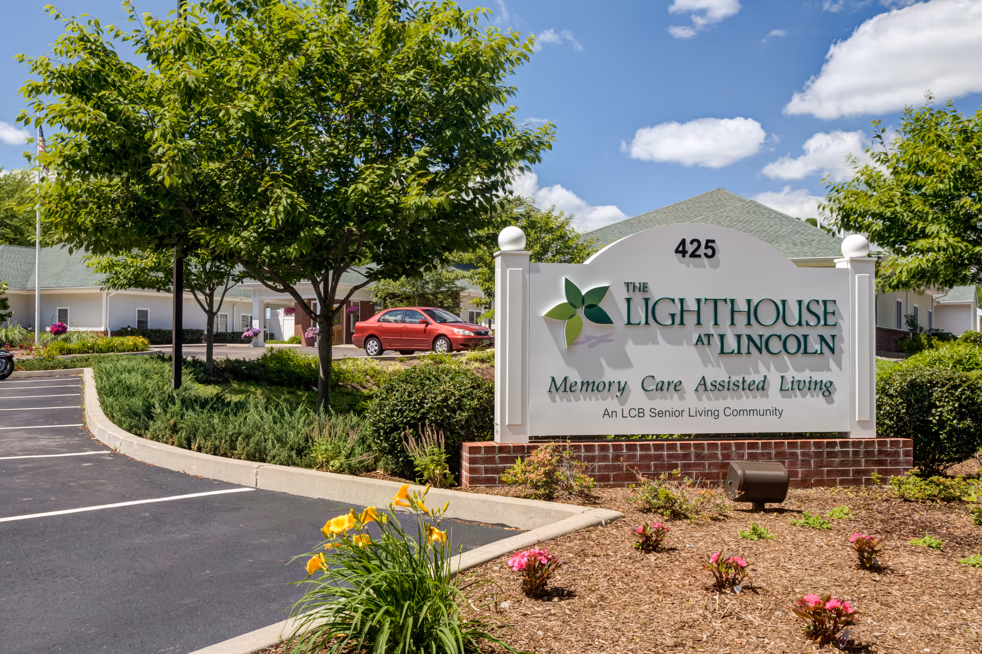 Outdoor view of the entrance sign for The Lighthouse at Lincoln, a memory care assisted living community, with a parking lot, trees, flowers, and a building in the background under a blue sky with clouds.